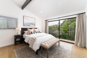 A bedroom with a large bed, a bench, a nightstand, a window with blinds, and a view of greenery.
