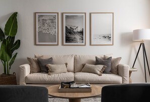 A living room with a beige sofa, three framed photographs on the wall, a floor lamp, and a coffee table with a book and decorative items.