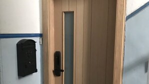 A wooden door with a vertical window and a black intercom on the wall beside it.
