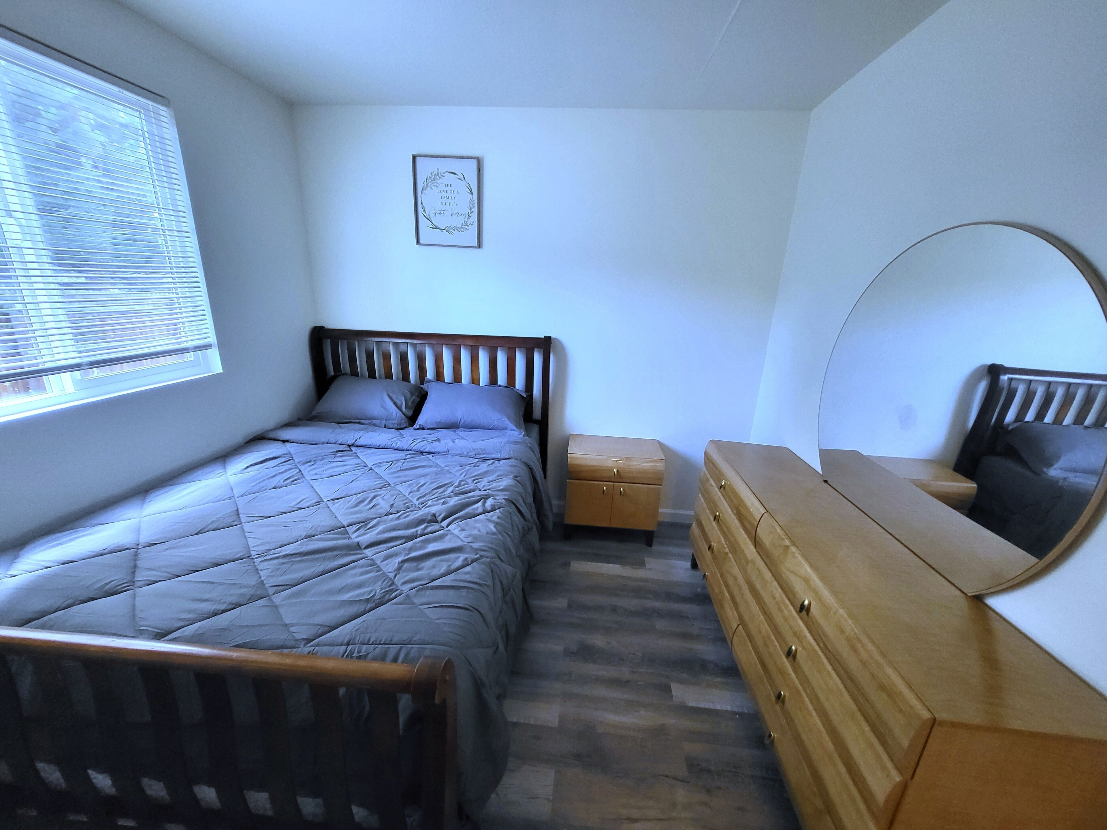 Ein Schlafzimmer mit einem Bett, einem Schminktisch, einem Nachttisch, einem Fenster mit Jalousien und einem Wandspiegel.
