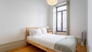 Un dormitorio minimalista con una cama de madera, ropa de cama blanca, una mesita de noche con una lámpara, una ventana con vistas a edificios y un calefactor portátil.
