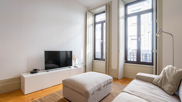 A modern living room with a flat-screen TV on a white console, a beige sofa, and a large window with a view of the street.