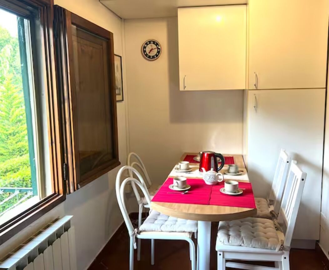 A dining area with a table set for four, white chairs, a clock on the wall, and a window with blinds.