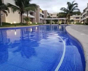 A swimming pool with blue tiles, surrounded by palm trees and a building with balconies. - Description (Mezcales)