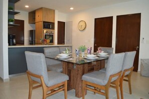 A dining area with a glass table surrounded by four grey chairs, wooden cabinets above, and a clock on the wall. - Description (Mezcales)
