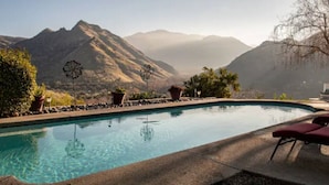 Piscina con montañas al fondo, rodeada de plantas en macetas y un cielo despejado.