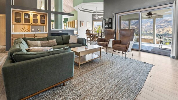 Amplia sala de estar con un sofá grande, una mesa de centro y una silla. Se ve el exterior a través de ventanas grandes.