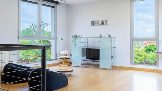 Ein moderner, gut beleuchteter Raum mit großen Fenstern, einem Bett, einem Fernseher und einem Schild mit der Aufschrift 'RELAX'.