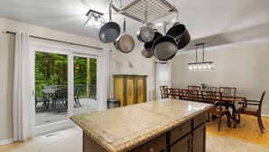 Une cuisine dotée d’un plan de travail en granit, de casseroles suspendues, d’un coin repas avec des chaises en bois et d’une vue sur une terrasse extérieure, accessible par une porte vitrée.