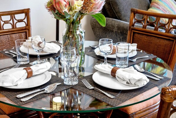 Une table à manger dressée pour six personnes, avec de la vaisselle en verre transparent, des assiettes blanches et des couverts en argent, entourée de chaises en osier.