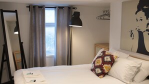 Ein ordentlich bezogenes Bett mit weißen Leinentüchern, einem gemusterten Kopfkissen und einem kleinen Nachttisch mit Lampe.