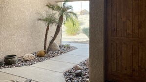 A modern entryway with a concrete floor, a wooden door, a palm tree, and a small garden area with rocks and a pot. - Sante Fe Style pool home (Lake Havasu City)