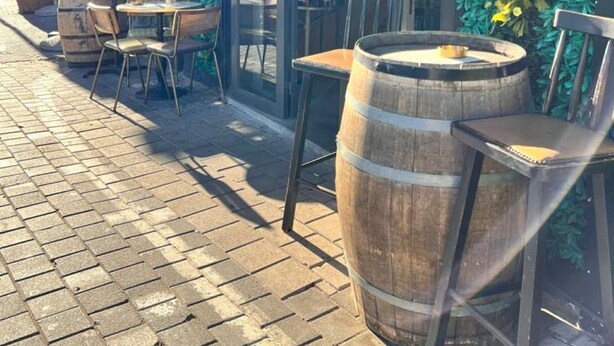 Un espacio al aire libre con sillas y mesas de madera, un barril grande, y un escaparate de vidrio donde se ven diversas plantas y botellas.