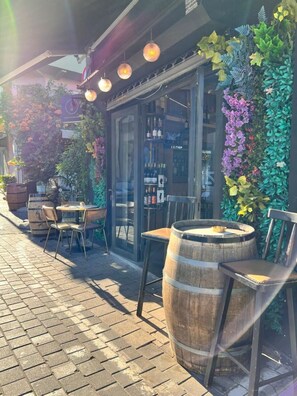 An outdoor seating area with wooden chairs and tables, a large barrel, and a glass storefront with a variety of plants and bottles visible inside.