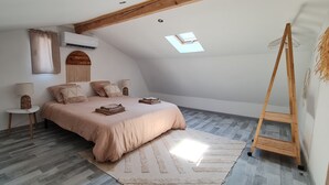 Un dormitorio con techo inclinado, una cama con colcha color beige, una mesita redonda pequeña con una lámpara y una escalera de madera.