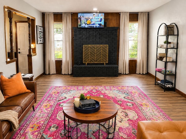 A living room with a black brick fireplace, a television mounted above it, a wooden coffee table with books and a candle, a colorful rug, and a leather sofa.
