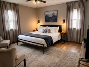 A bedroom with a bed, bedside lamps, a ceiling fan, a painting, and a television. - Discover Enid - Centrally Located Beautiful Brick Home (Enid)