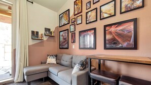 A modern living room with a grey sectional sofa, a wooden table, and a bar stool. The room features a large window with a view of the outside. - River Rock Studio w/private courtyard, 20 minutes to Zion (Virgin)