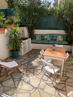 A patio with a wooden table and white chairs, a green sofa with tropical cushions, and a potted plant.
