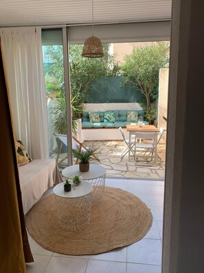 A modern living room with a glass door leading to an outdoor patio. The room features a round table with a woven rug, a sofa with cushions, and a hanging light fixture.