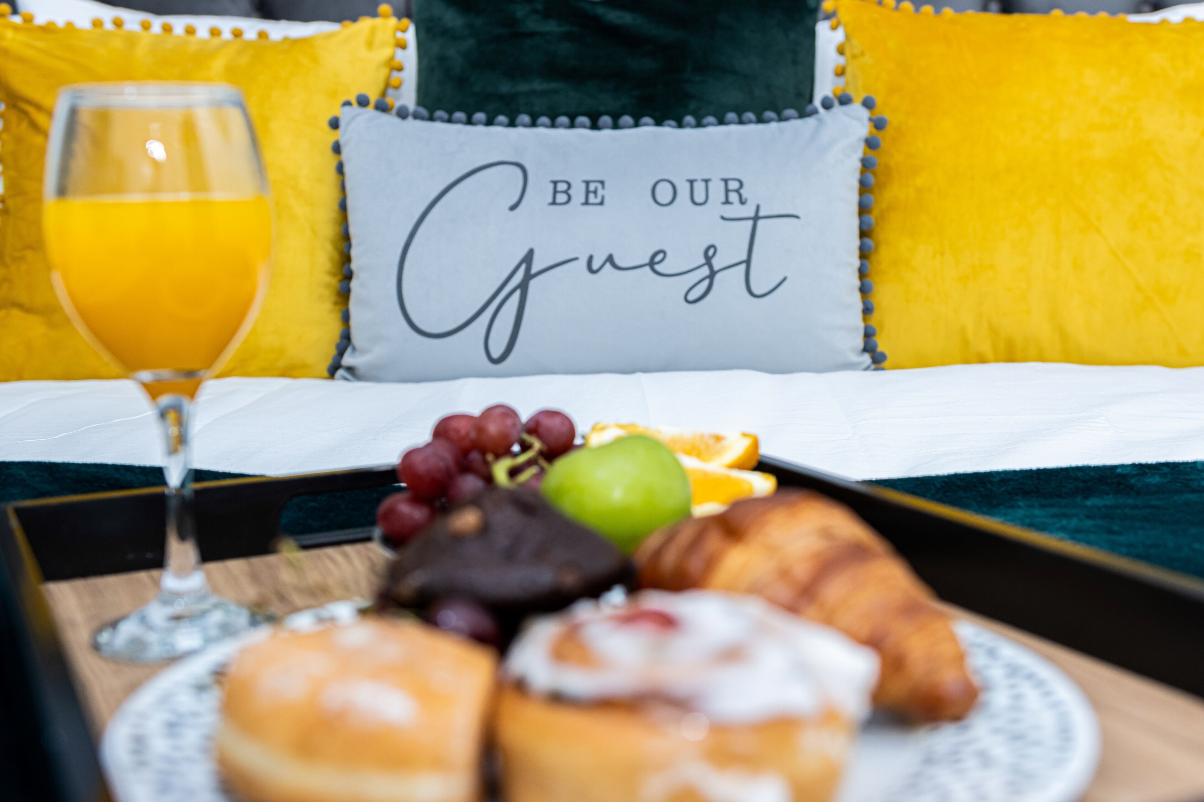 A breakfast tray with croissants, fruits, and a glass of orange juice on a bed with a pillow that says "BE OUR Guest".