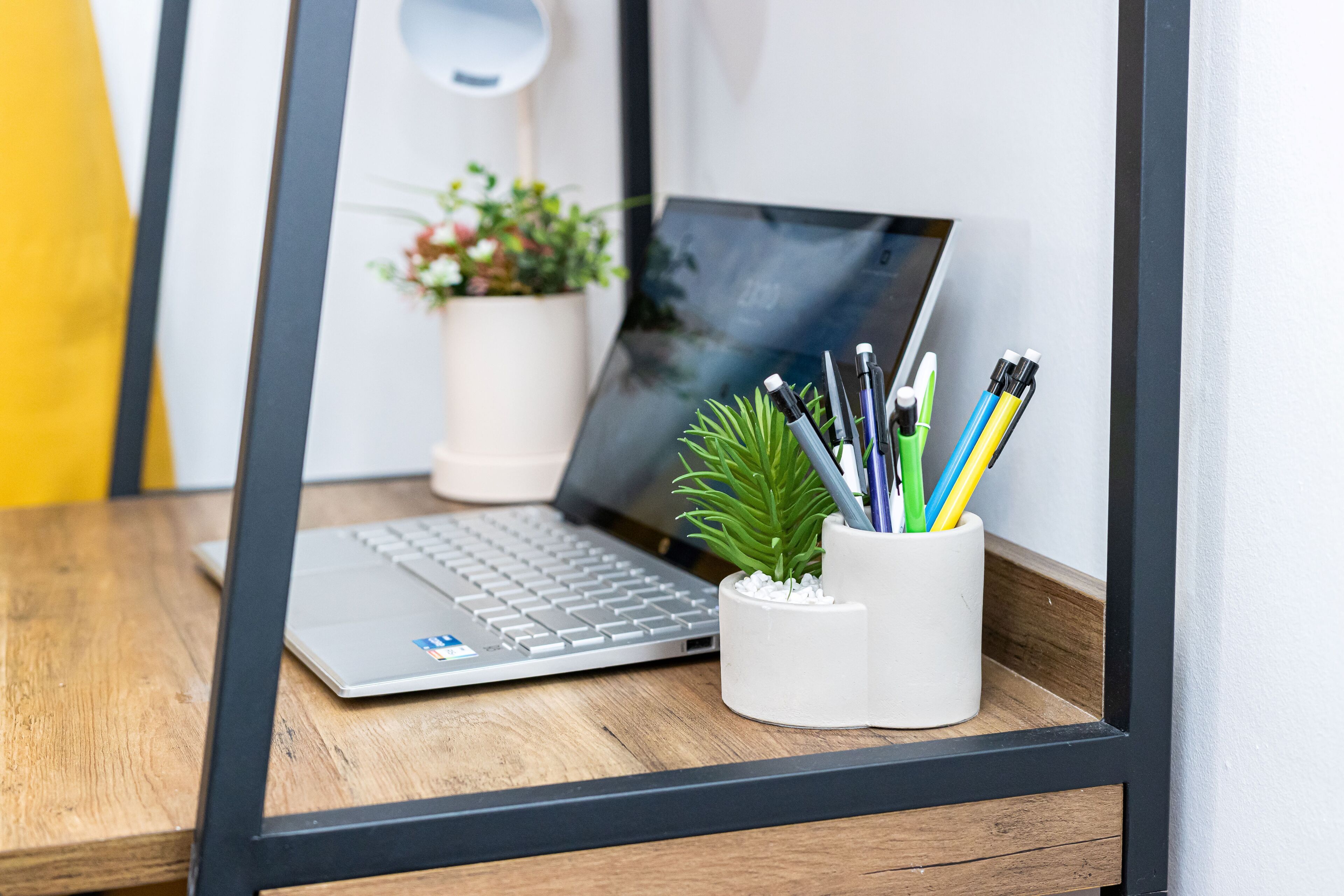 A laptop on a wooden desk with a white cup holding pens and pencils.