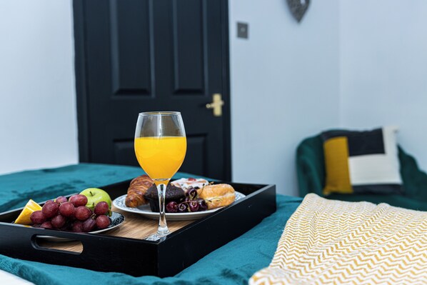 A breakfast tray with fruits, a glass of orange juice, and pastries on a bed with a teal bedspread and a patterned pillow. - Cosy 2bedroom House-perfect for Long & Short Stay (Wolverhampton)