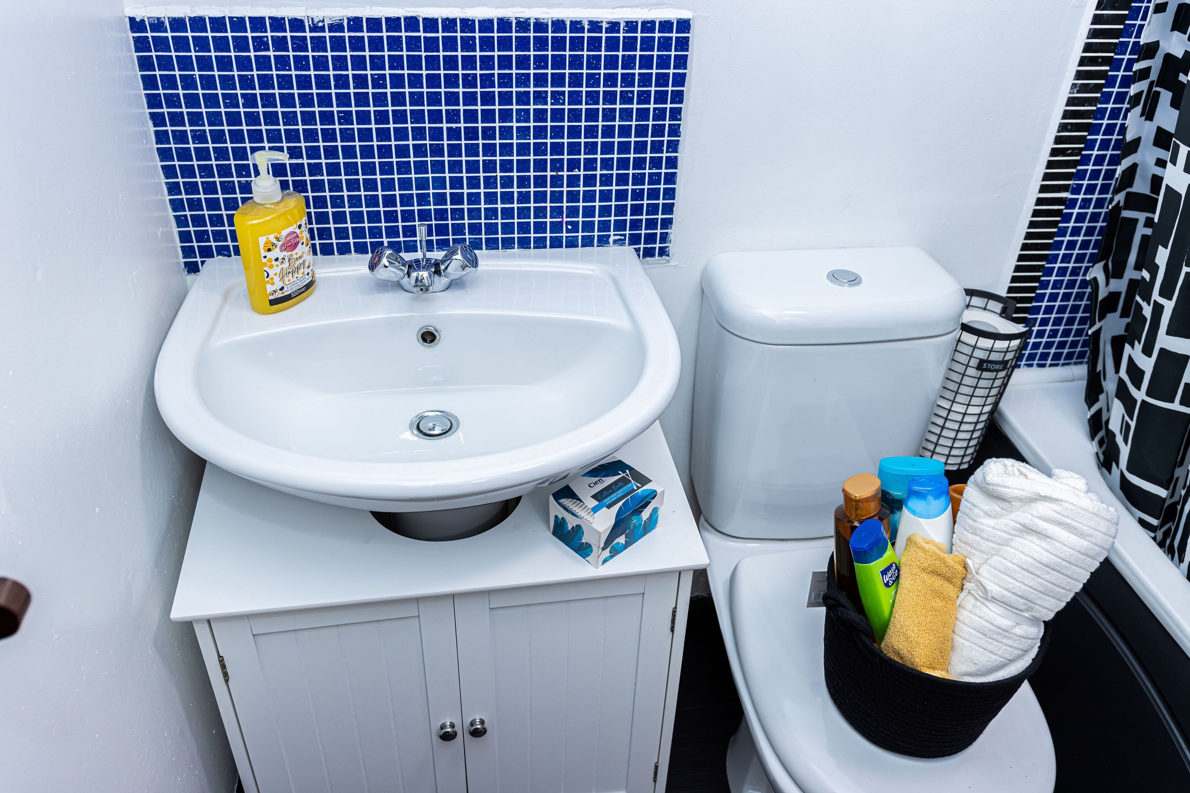 A bathroom with a white sink, blue tiled wall, and a toilet with a basket of toiletries.