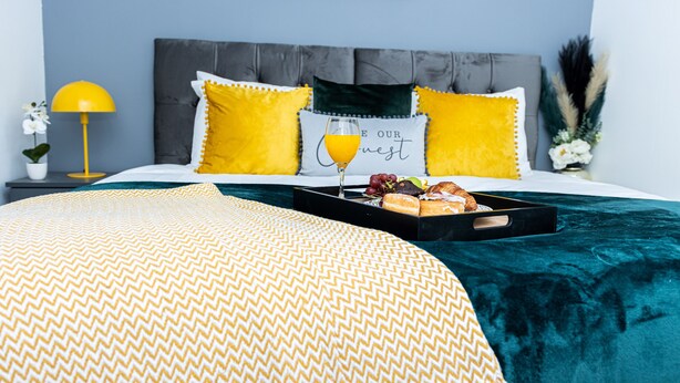 A neatly made bed with a yellow and white patterned bedspread, a tray with breakfast items, and a glass of orange juice on the bed. - Cosy 2bedroom House-perfect for Long & Short Stay (Wolverhampton)