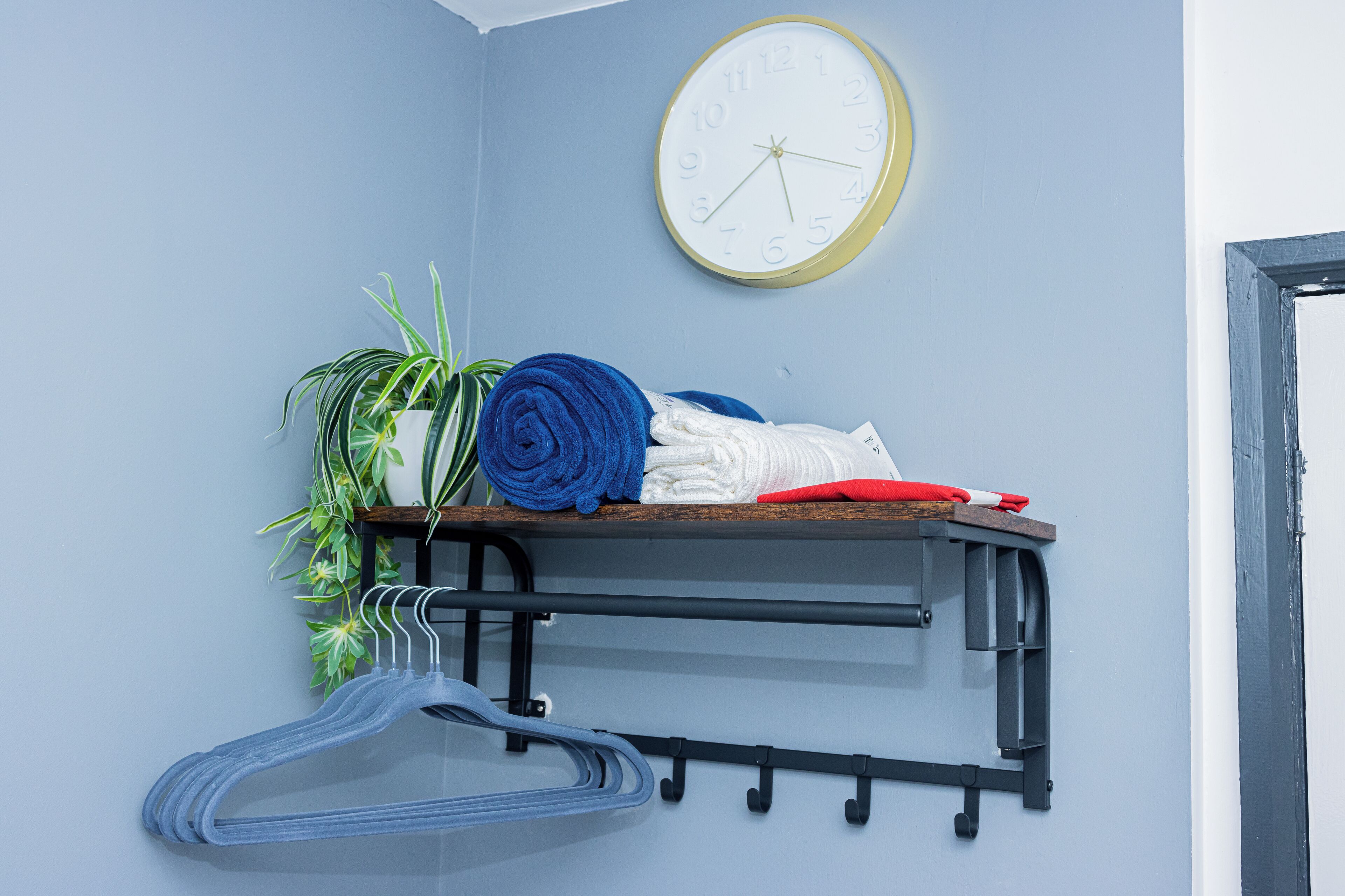 A corner of a room with a wall-mounted clock, a shelf with folded towels, a plant, and a clothes rack.