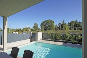 A swimming pool with two wicker chairs, a covered patio area, and a view of a residential neighborhood with houses and trees.