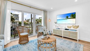 Amplia sala de estar con un gran ventanal, muebles de mimbre, una mesa de centro redonda y un televisor montado que muestra una escena de playa.