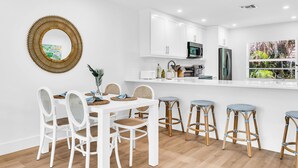 Una cocina moderna con armarios blancos, un microondas y un refrigerador. Hay una mesa de comedor con sillas blancas y una zona de taburetes de bar.