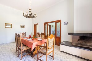 Comedor con piso de mármol, sillas de madera, mesa con platos y vasos, una araña y una chimenea.