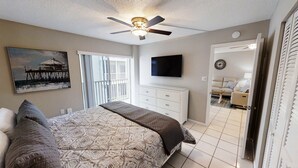 A bedroom with a bed, a ceiling fan, a TV, a painting, and a balcony.