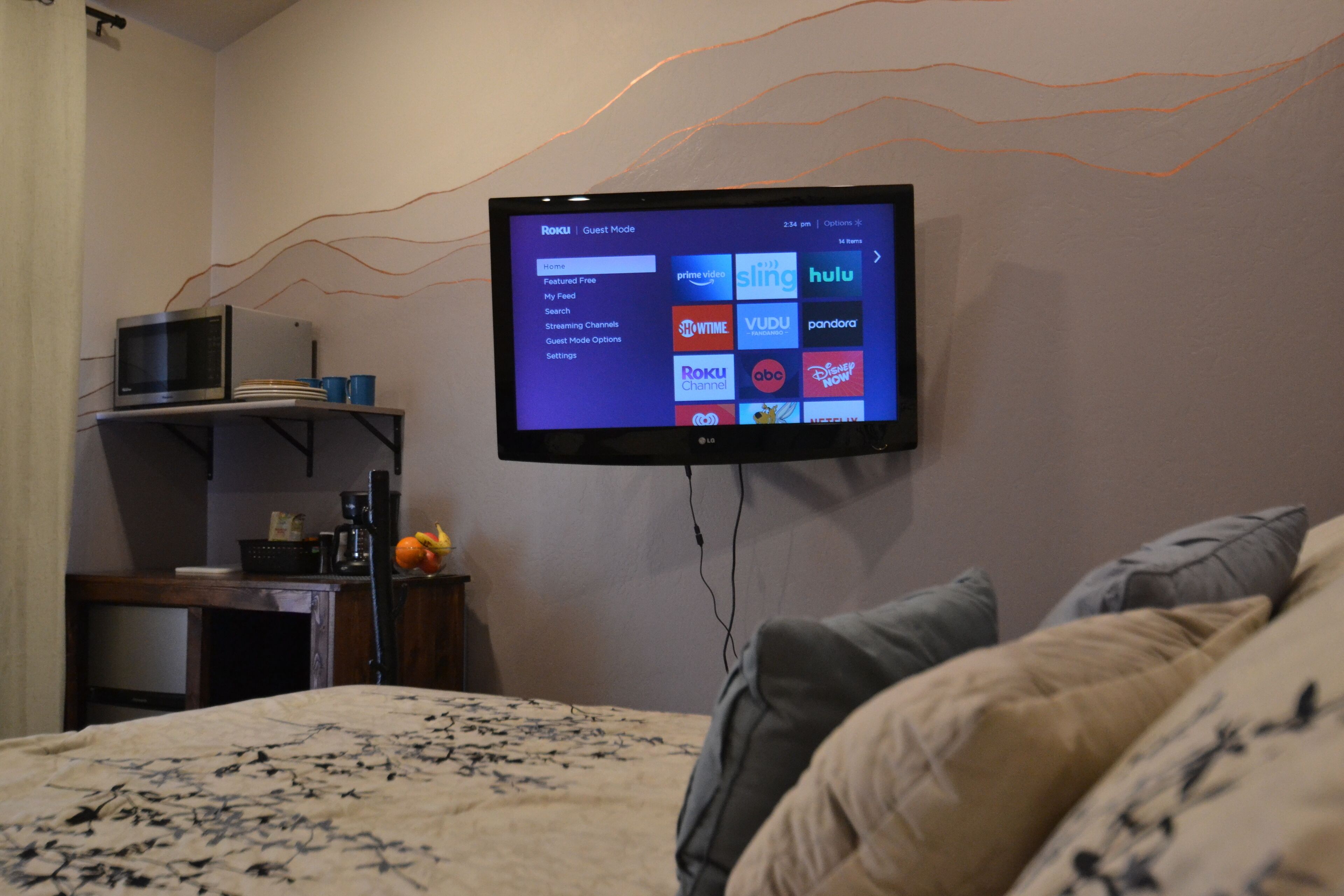 A bedroom with a bed, a TV mounted on the wall displaying a streaming service interface, a microwave on a shelf, and a small table with a fruit bowl.