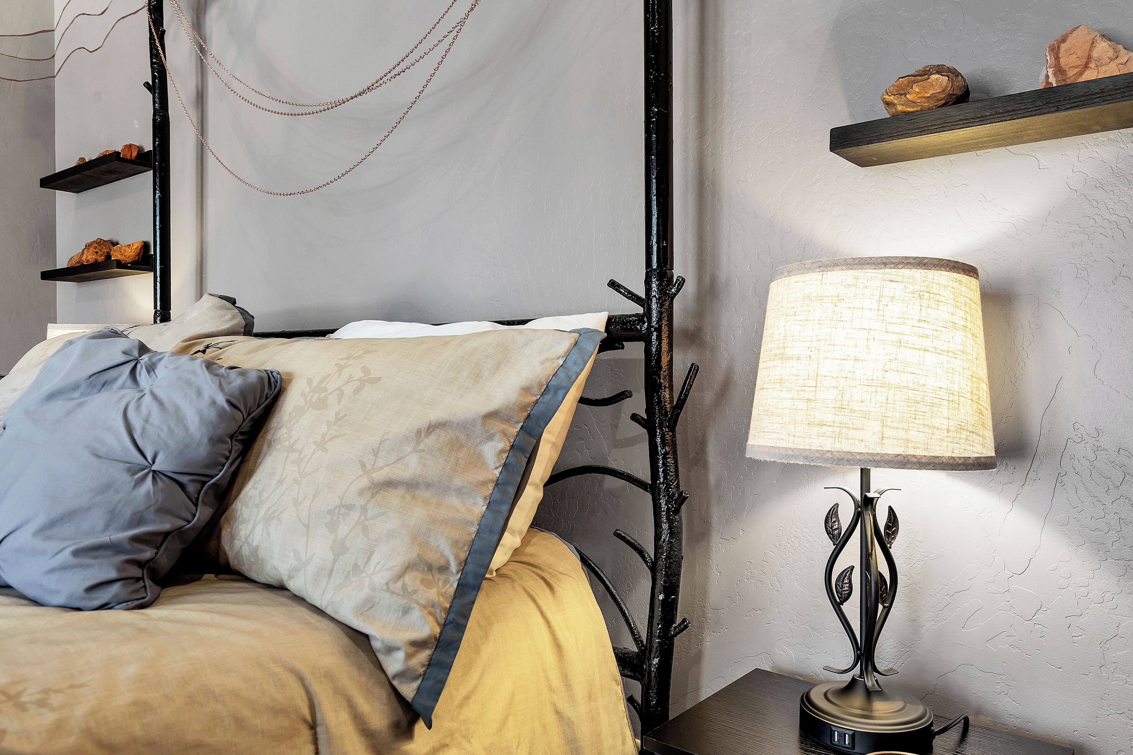 Una cama bien hecha con un cojín estampado y uno azul decorativo, una mesita de noche con una lámpara, y un estante con adornos.
