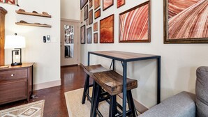Un salon moderne avec un banc en bois, une table et un canapé. Plusieurs œuvres d’art encadrées ornent le mur, et une lampe est posée sur une table d’appoint.