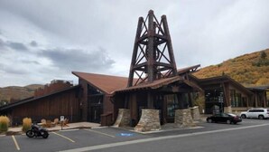 Un edificio rústico con una destacada estructura metálica en torre, cimientos de piedra y un estacionamiento con algunos vehículos.