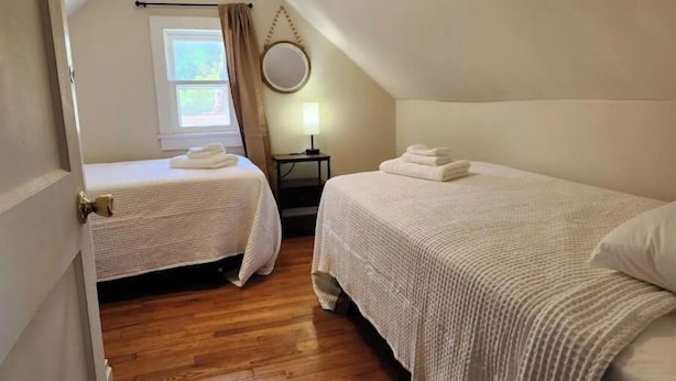Una habitación con dos camas, una ventana con cortinas, una mesita con lámpara y un espejo redondo en la pared.