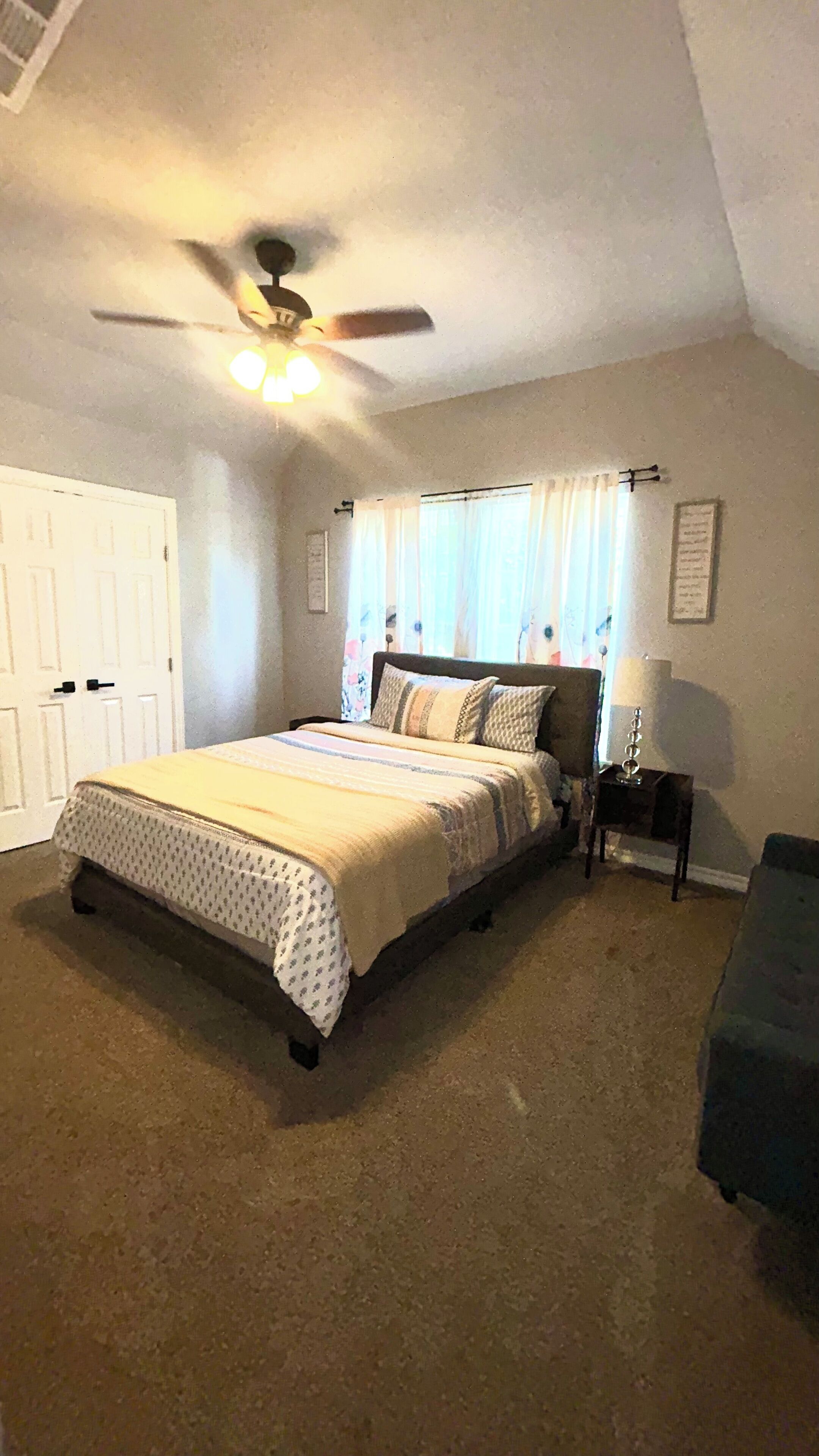 Une chambre à coucher avec un lit, un ventilateur de plafond, une table de chevet, une lampe et une fenêtre avec des rideaux.