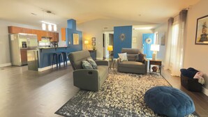 Ein modernes Wohnzimmer mit einer Couch, einem Couchtisch und einem Teppich. Es gibt eine Küchenzeile mit einem Kühlschrank und Barhocker.