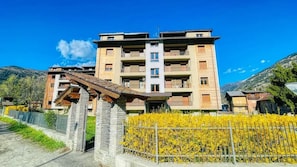 Un edificio de varios pisos con balcones, una entrada de piedra y un seto amarillo lleno de flores.