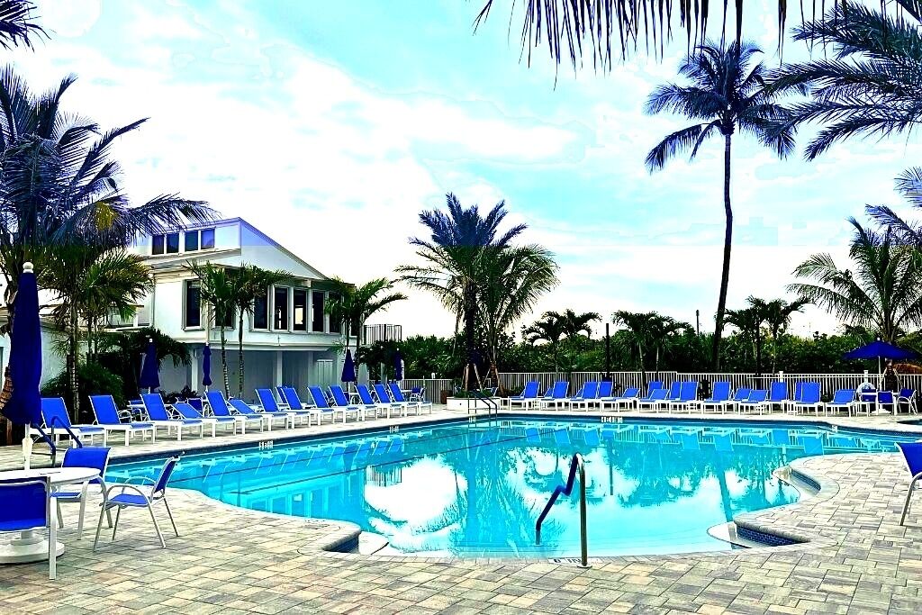A swimming pool with blue chairs and palm trees, surrounded by a building with a white facade and a balcony.