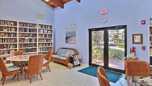 A cozy library with wicker furniture, bookshelves, and a large window with a view of greenery. - Private Beach Resort Condo, Hutchinson Island, FL (Fort Pierce)
