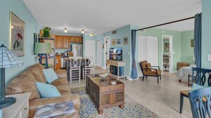 A living room with a brown sofa, a coffee table, and a TV. There is a kitchen in the background with wooden cabinets and a dining area with chairs. - Private Beach Resort Condo, Hutchinson Island, FL (Fort Pierce)