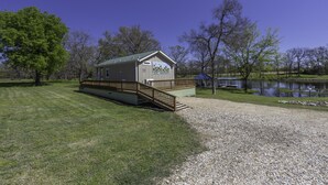 Eine kleine Holzhütte mit einer Holzterrasse und einem Kiesweg, umgeben von Bäumen und einem Gewässer.