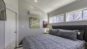 Ein Schlafzimmer mit einem großen Bett, einem Nachttisch, einem an der Wand befestigten Fernseher und einem Fenster mit Blick auf Bäume.