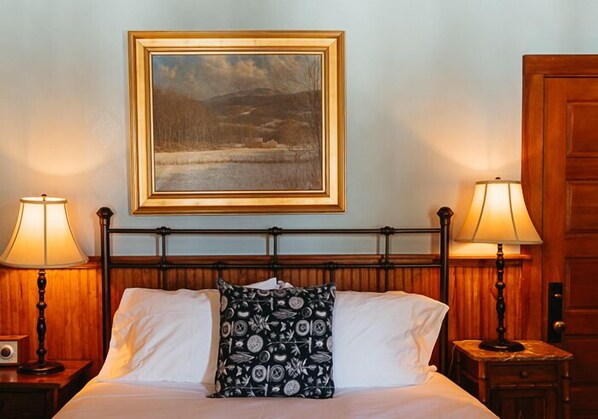 A hotel room with a bed, two bedside lamps, a framed painting above the bed, and a wooden door. - Deer Mountain Inn (Haines Falls)
