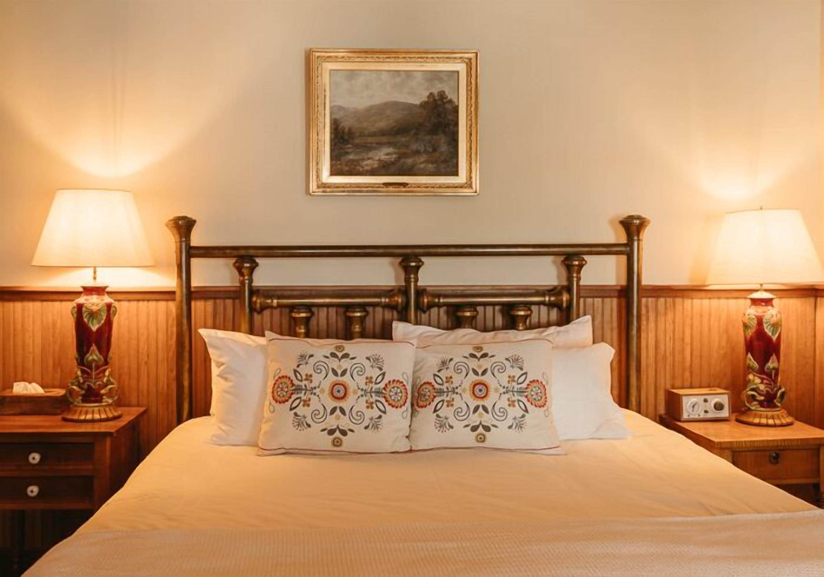 A neatly made bed with decorative pillows, a framed landscape painting above, and two bedside lamps on wooden nightstands.